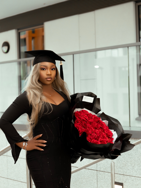 A woman in a graduation cap and black dress holding a massive bouquet of red roses indoors. — portrait photography London by Lamss Media